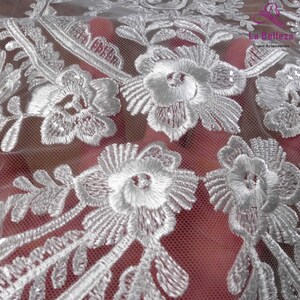 K&ouml;nnte beinhalten: Wei&szlig;er Blumenspitzenstoff mit einem zarten Muster. Die Spitze ist durchsichtig und hat eine weiche, flie&szlig;ende Textur. La Belleza Gown Accessories ist auf den Stoff gedruckt.