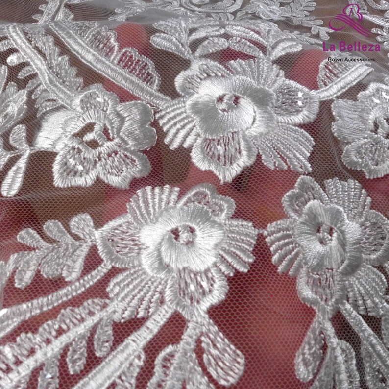 K&ouml;nnte beinhalten: Wei&szlig;er Blumenspitzenstoff mit einem zarten Muster. Die Spitze ist durchsichtig und hat eine weiche, flie&szlig;ende Textur. La Belleza Gown Accessories ist auf den Stoff gedruckt.