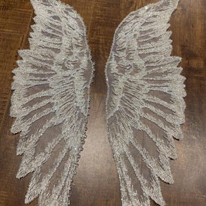 May include: A pair of white angel wings made of sheer fabric with a beaded design. The wings are delicate and intricate, with a shimmering effect.