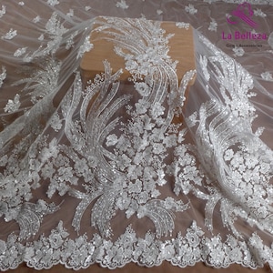 May include: White tulle fabric with silver glitter and floral embroidery. The fabric has a delicate, flowing texture and is perfect for adding a touch of elegance to any project.