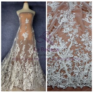 May include: White beaded lace fabric with a floral design. The fabric is sheer and has a delicate texture. The design features small flowers and leaves, as well as larger, more elaborate floral motifs. The fabric is perfect for creating wedding gowns, evening dresses, and other special occasion garments.