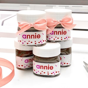 May include: Four small glass jars with white lids and pink ribbon bows. Each jar has a brown chocolate spread inside and a label that says "annie" with pink hearts.