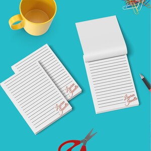 Może przedstawiać: Dwa białe notesy w linie z imieniem "Lauren Janelle" napisanym czerwonym, ozdobnym pismem w prawym dolnym rogu każdego notesu. Notesy leżą na niebieskiej powierzchni z żółtym kubkiem, czerwonymi nożyczkami i długopisem.