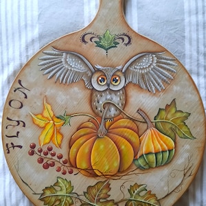Può includere: Un tagliere rotondo in legno con un design dipinto di un gufo appollaiato su una zucca e una vite con foglie. Il tagliere ha un manico e le parole "Fly On" e "Liza Design" sono dipinte sulla superficie.