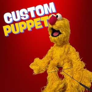 May include: A yellow custom puppet with a red nose and mouth open wide. The puppet is made of fuzzy material and has long arms and legs. The puppet is being held by a black rod.