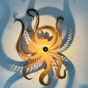 Puede incluir: Una lámpara de pared decorativa con forma de pulpo. La lámpara está hecha de un material marrón claro con detalles recortados y una fuente de luz central. La luz proyecta una sombra del pulpo en la pared. La lámpara tiene una gema azul oscuro en el centro.