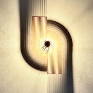 Può includere: Un'applique da parete moderna con una luce calda. L'apparecchio presenta un pannello rettangolare verticale con linee verticali e un elemento curvo e circolare. L'applique proietta un'ombra sul muro.