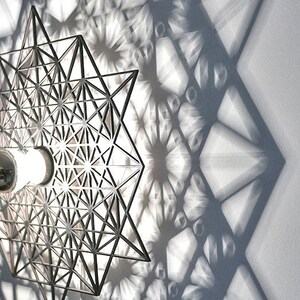 May include: A silver metal wall sconce with a geometric design casts a shadow of repeating geometric shapes on a white wall. The sconce has a light bulb in the center.