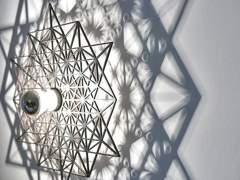 May include: A silver metal wall sconce with a geometric design casts a shadow of repeating geometric shapes on a white wall. The sconce has a light bulb in the center.