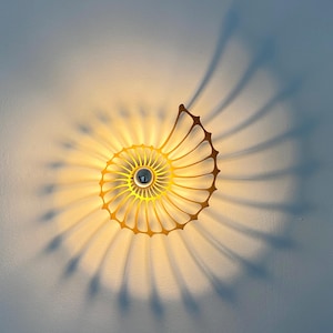 Puede incluir: Un aplique de pared con forma de concha de nautilo y marco de metal dorado. La lámpara emite una luz cálida y brillante desde el centro, proyectando largas sombras radiantes que imitan el patrón en espiral de la concha sobre la pared.
