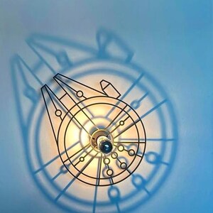 Könnte beinhalten: Eine schwarze Metall-Wandleuchte in Form des Raumschiffs Millennium Falcon. Das Licht wirft einen Schatten auf die hellblaue Wand und verstärkt das Design. Die Leuchte hat eine zentrale Glühbirne.
