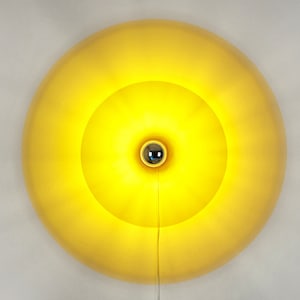 Könnte beinhalten: Eine leuchtend gelbe, kreisförmige Wandleuchte. Das Design weist konzentrische Kreise mit einer zentralen, metallischen Glühbirne auf. Ein dünnes, weißes Kabel hängt von unten. Das Licht ist an und erzeugt einen warmen Schein.