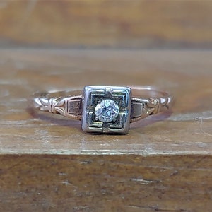May include: A vintage engagement ring with a single round diamond set in a square frame. The ring is made of two-toned gold, with a white gold frame and a rose gold band.