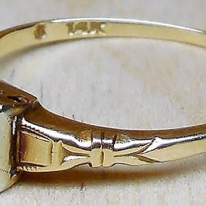 May include: A gold ring with a single diamond set in a square bezel. The ring has a delicate engraved design on the band. The ring is marked "14K".