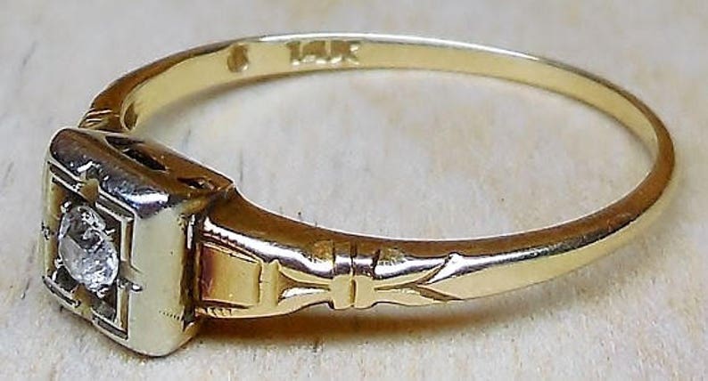 May include: A gold ring with a single diamond set in a square bezel. The ring has a delicate engraved design on the band. The ring is marked "14K".