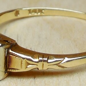 May include: A gold ring with a square-shaped setting featuring a small diamond. The band is engraved with a delicate floral design and is marked "14K".