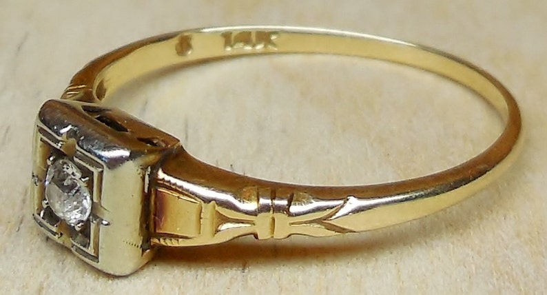 May include: A gold ring with a square-shaped setting featuring a small diamond. The band is engraved with a delicate floral design and is marked "14K".