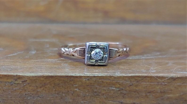 May include: A vintage engagement ring with a single round diamond set in a square frame. The ring is made of two-toned gold, with a white gold frame and a rose gold band.