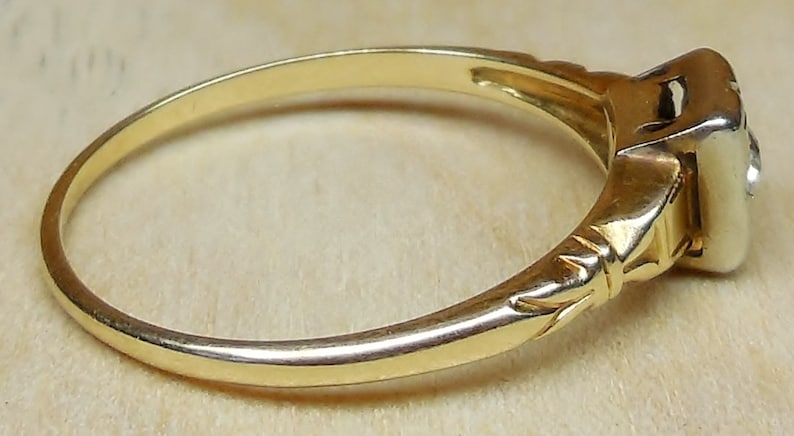 May include: A gold ring with a single diamond set in a square-shaped prong setting. The band is plain and has a delicate, engraved design.