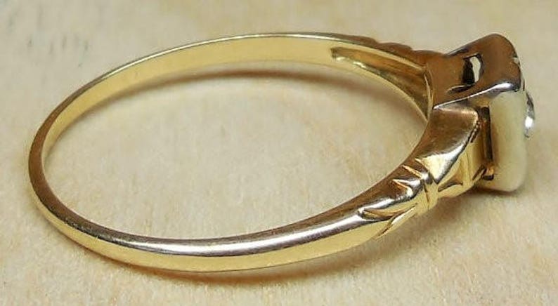 May include: A gold ring with a single diamond set in a square bezel. The ring has a delicate band with a decorative design.