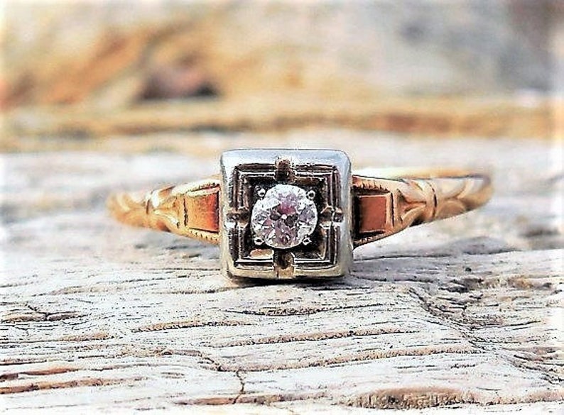 May include: A vintage engagement ring with a single round diamond set in a square platinum setting. The band is yellow gold and has a delicate, ornate design.