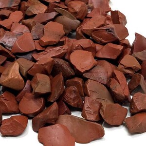 May include: A pile of red jasper chips, a type of gemstone, with a variety of shapes and sizes. The chips are a deep red color with some lighter brown and white streaks.