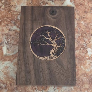 May include: A rectangular dark brown wooden block featuring a circular inlay. The inlay consists of a deep purple resin background with a light-colored wood cutout of a tree with intricate, sprawling branches and a textured root system.