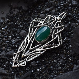 May include: A silver pendant with a green gemstone set in a wire-wrapped design. The pendant is on a black background.