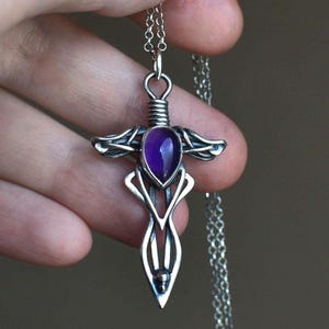 May include: A silver-toned pendant necklace featuring a dagger-shaped design. The pendant has a teardrop-shaped, deep purple gemstone at its center. The intricate metalwork includes stylized wings and decorative flourishes, suspended from a delicate chain.