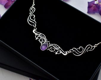 May include: A silver-toned necklace featuring intricate, swirling metalwork patterns and a single, central teardrop-shaped purple gemstone. The necklace is displayed inside a black rectangular box, with delicate chain links extending from both sides.