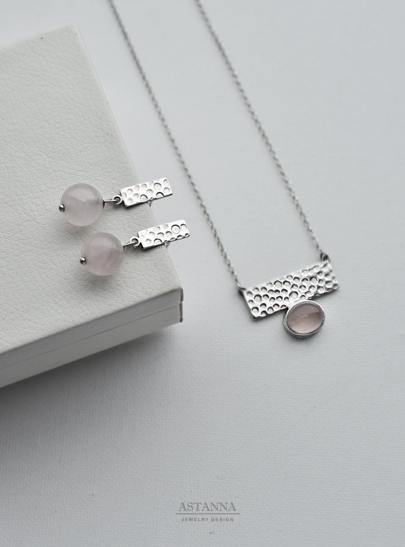 May include: A silver necklace and earrings set with a hammered metal design and pink rose quartz stones. The necklace features a rectangular pendant with a rose quartz stone in the center. The earrings are drop earrings with a rose quartz stone and a hammered metal bar.
