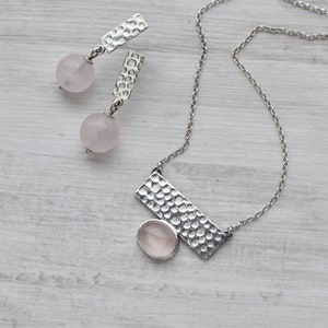 May include: A silver necklace and earrings set with a hammered metal design and pink rose quartz stones. The necklace features a rectangular pendant with a larger rose quartz stone.