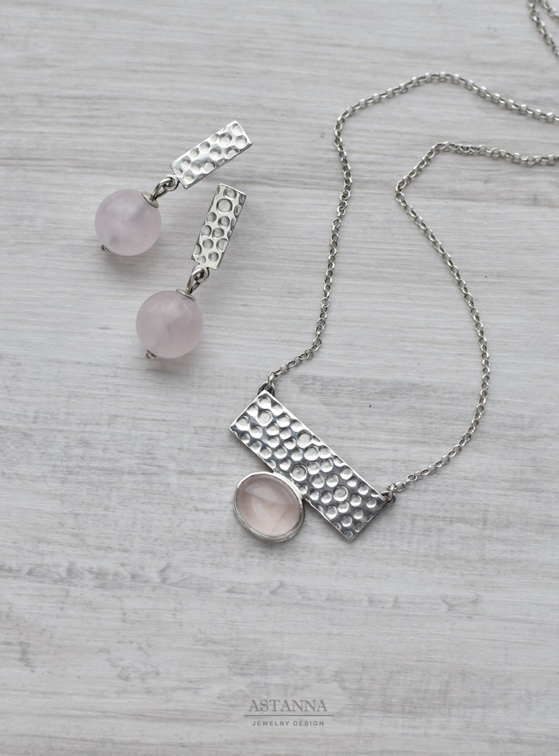 May include: A silver necklace and earrings set with a hammered metal design and pink rose quartz stones. The necklace features a rectangular pendant with a larger rose quartz stone.
