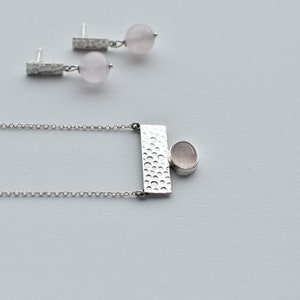 May include: A silver necklace with a textured rectangular pendant and a round pink gemstone. Matching earrings with a textured bar and a round pink gemstone.