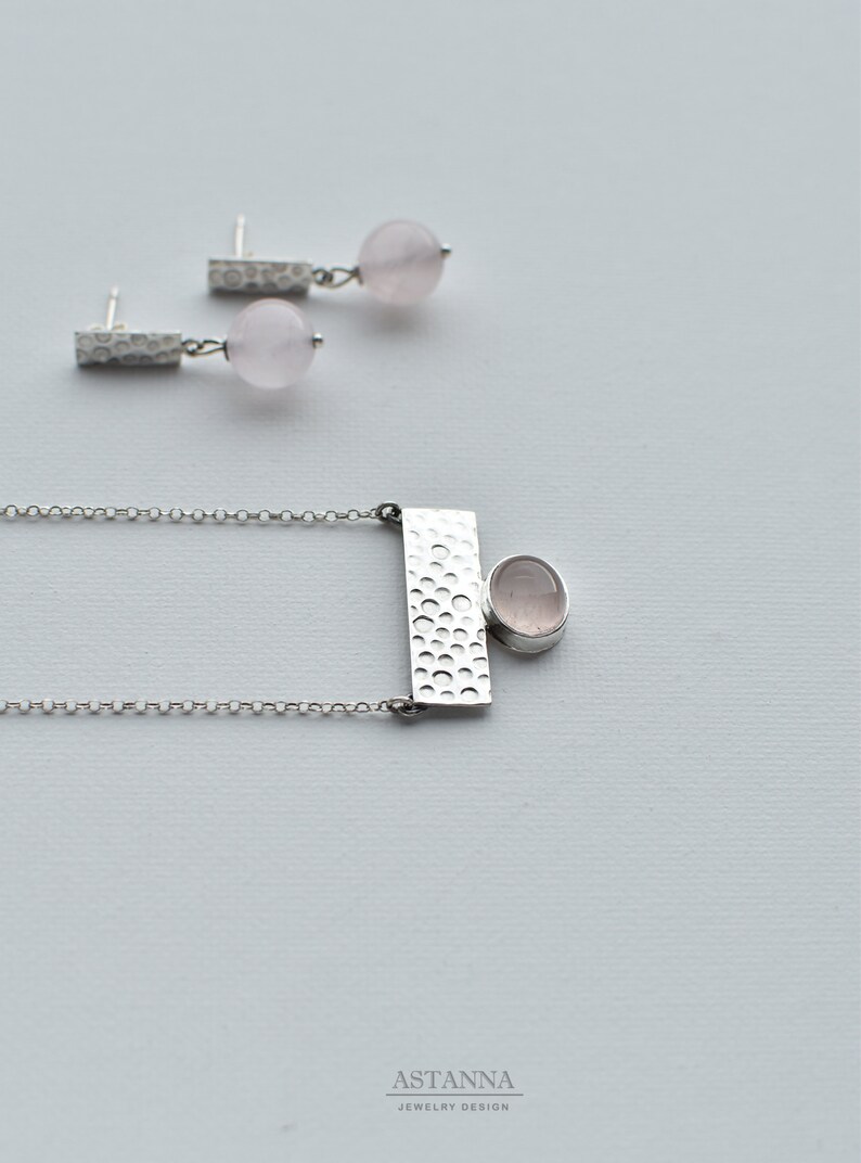 May include: A silver necklace with a textured rectangular pendant and a round pink gemstone. Matching earrings with a textured bar and a round pink gemstone.