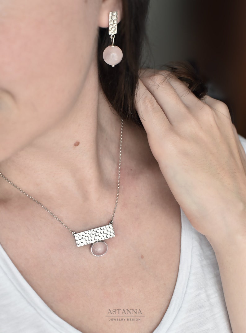 May include: A silver necklace with a rectangular pendant featuring a textured design and a pink gemstone. The necklace is paired with matching earrings with a similar design and pink gemstones.