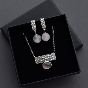 May include: A silver necklace and earrings set with a textured bar pendant and round pink stones. The jewelry is displayed in a black box with the brand name "ASTANNA JEWELRY DESIGN" printed on the bottom.