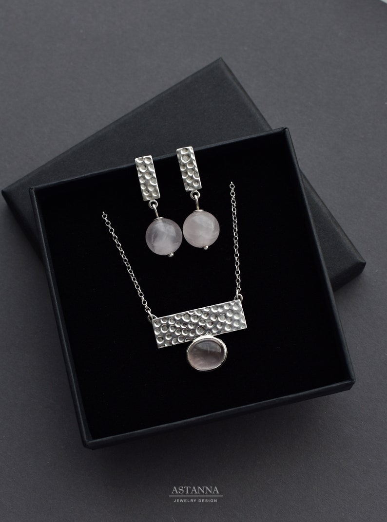 May include: A silver necklace and earrings set with a textured bar pendant and round pink stones. The jewelry is displayed in a black box with the brand name "ASTANNA JEWELRY DESIGN" printed on the bottom.