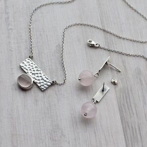 May include: A silver necklace with a hammered rectangular pendant and a pink oval gemstone. Matching earrings feature a pink round gemstone and a silver bar.