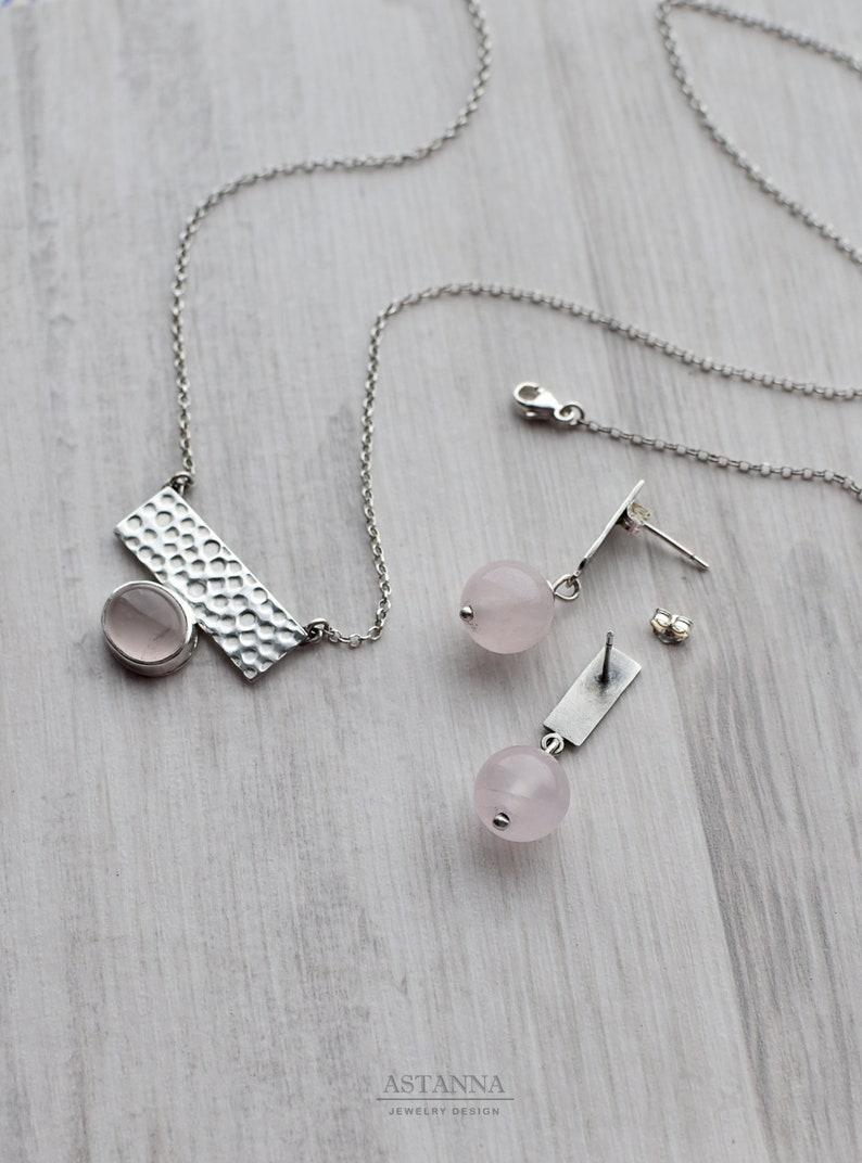 May include: A silver necklace with a hammered rectangular pendant and a pink oval gemstone. Matching earrings feature a pink round gemstone and a silver bar.