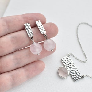 May include: A silver necklace and earrings set with rose quartz stones. The necklace features a hammered silver pendant with a rose quartz stone. The earrings are silver with hammered rectangular pendants and rose quartz stones.