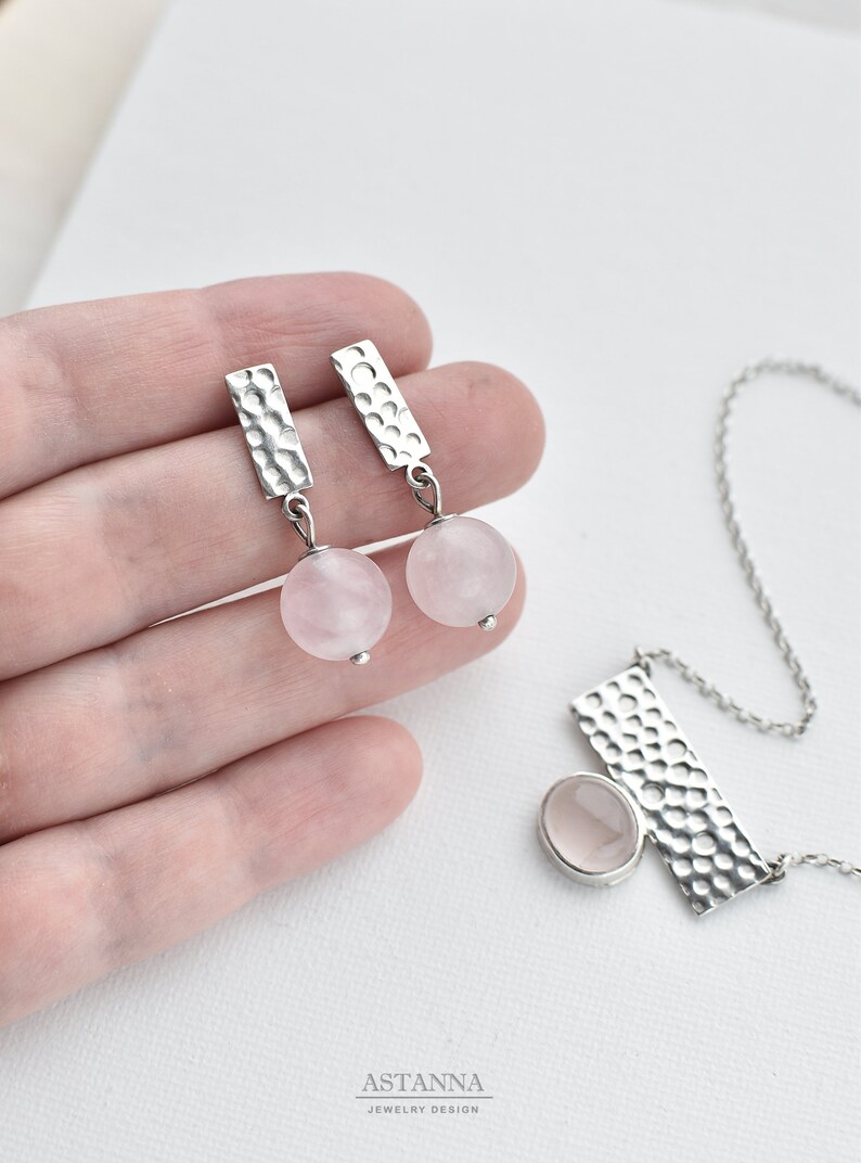 May include: A silver necklace and earrings set with rose quartz stones. The necklace features a hammered silver pendant with a rose quartz stone. The earrings are silver with hammered rectangular pendants and rose quartz stones.