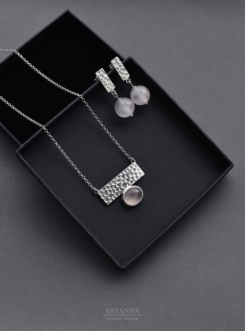May include: A silver necklace and earrings set with a textured bar pendant and round pink quartz stones. The jewelry is displayed in a black box.
