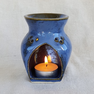 May include: A blue ceramic oil diffuser with a small candle inside. The diffuser has a decorative design with holes around the top and a wide opening in the front for the candle.