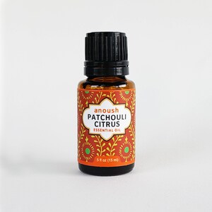 May include: A small amber glass bottle with a black cap, containing Patchouli Citrus essential oil. The label is orange and yellow with a green and white floral pattern. It reads "anoush PATCHOULI CITRUS ESSENTIAL OIL 5 fl oz (15 ml)"