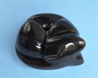 May include: A black ceramic incense burner shaped like a sleeping cat. The burner has a small hole on the top for incense sticks.
