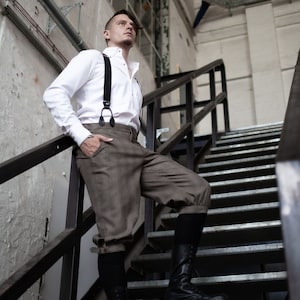 Puede incluir: Un hombre con una camisa blanca abotonada con tirantes y pantalones marrones se encuentra en una escalera de metal. Lleva botas negras y mira hacia arriba.