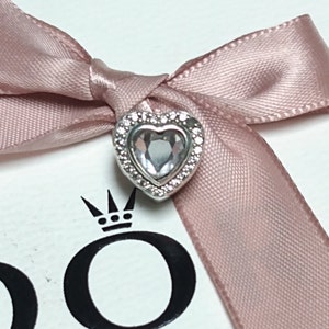 Puede incluir: Un charm de plata con un corazón de cristal transparente rodeado por un halo de cristales más pequeños. El charm está sobre una cinta rosa y una caja blanca con el logotipo de Pandora.