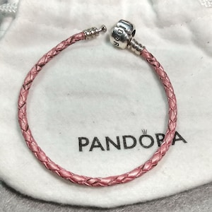 May include: A pink braided leather bracelet with a silver clasp. The bracelet is open, revealing the clasp mechanism. The word "PANDORA" is printed on a white bag in the background.