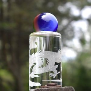 May include: A clear glass bottle stopper with an etched dragon design. The stopper is topped with a blue and red glass orb. The dragon is intricately detailed, and the overall aesthetic is elegant and artistic.